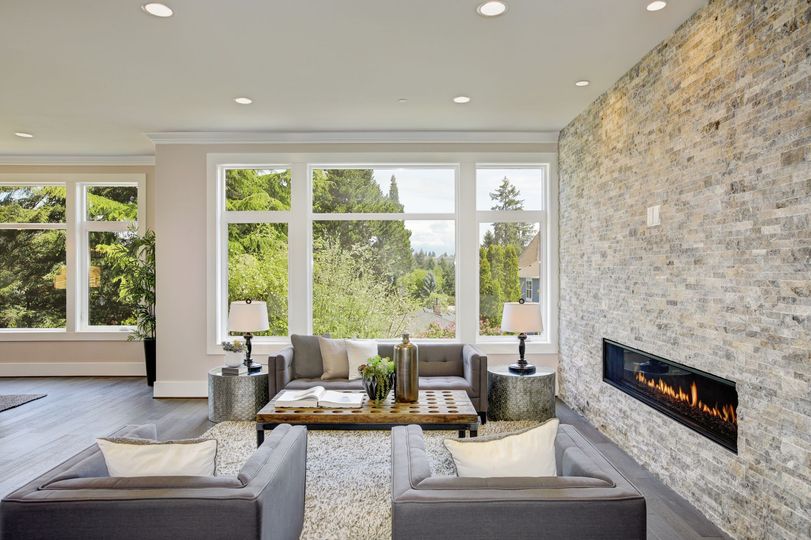Modern living room with stone fireplace, gray seating, and large windows overlooking trees