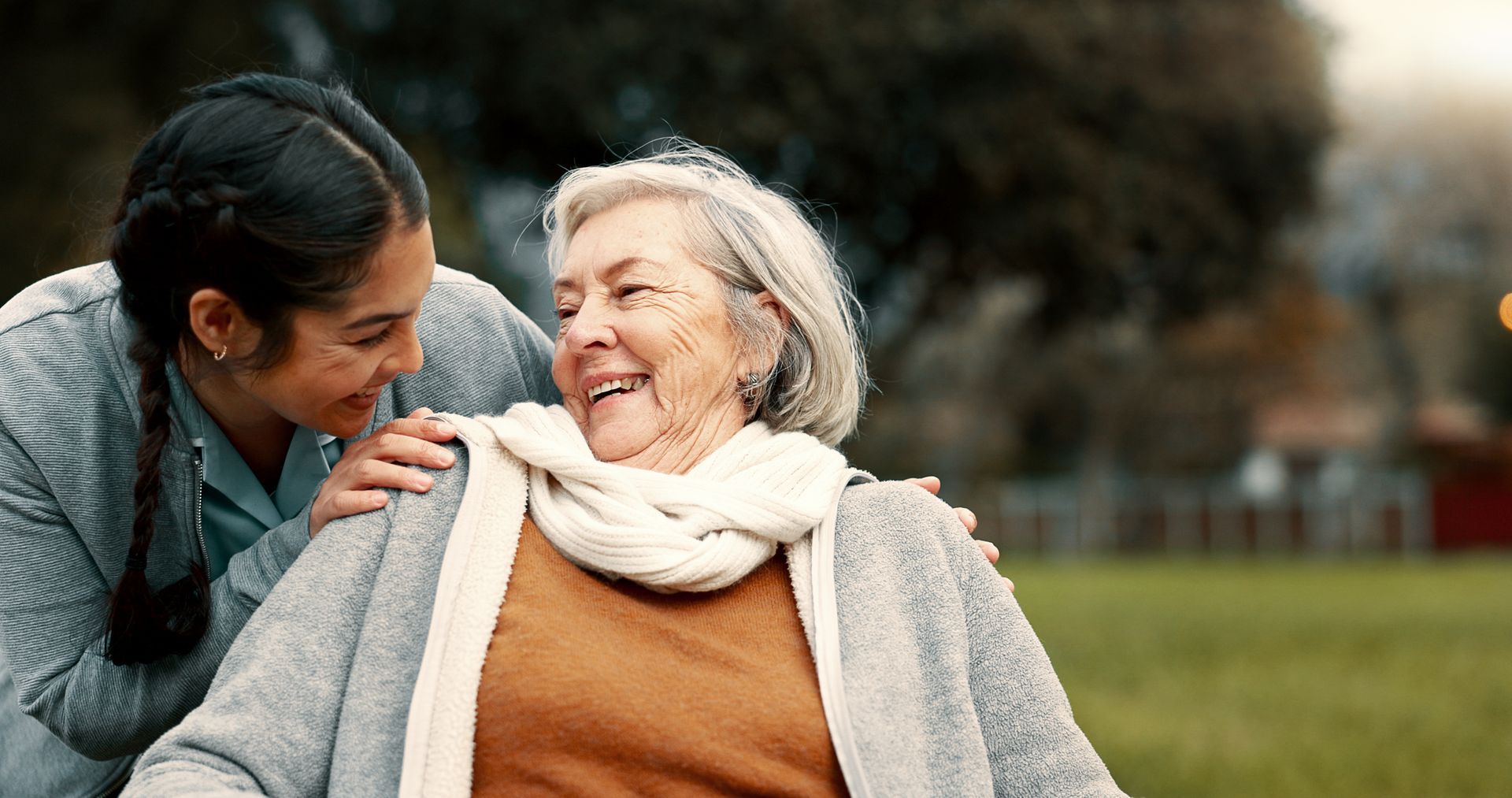 Person assisting an older adult outdoors, both smiling and interacting in a park setting. Person assisting an older adult outdoors, both smiling and interacting in a park setting.