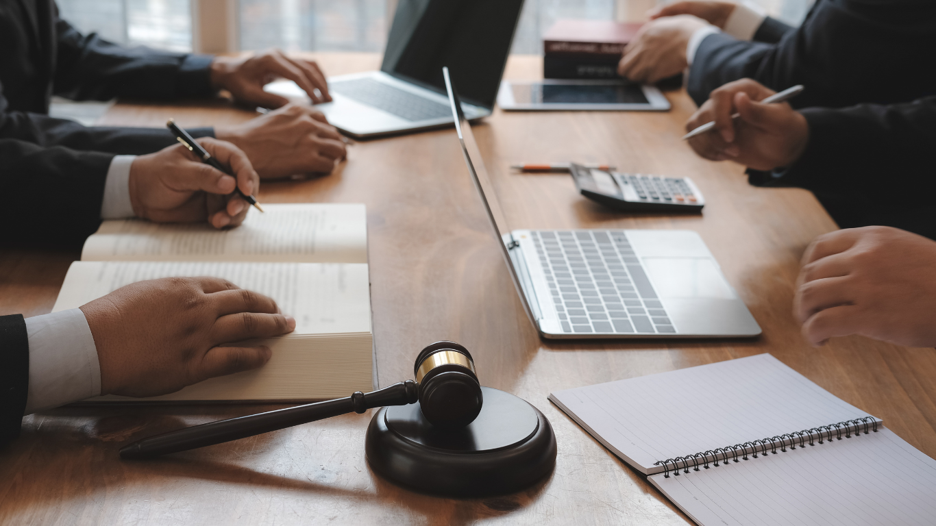 Businesspeople at a table, laptops, papers, gavel, and calculator present.
