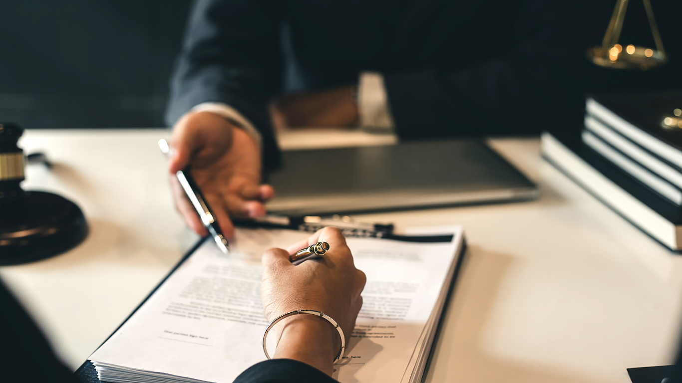 Person signing a document, pen in hand, as another person gestures towards it. Office setting.
