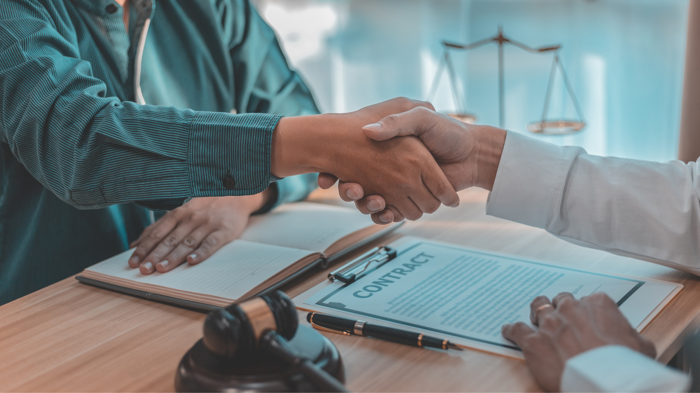 Two people shaking hands over a contract on a desk, scales of justice in background.