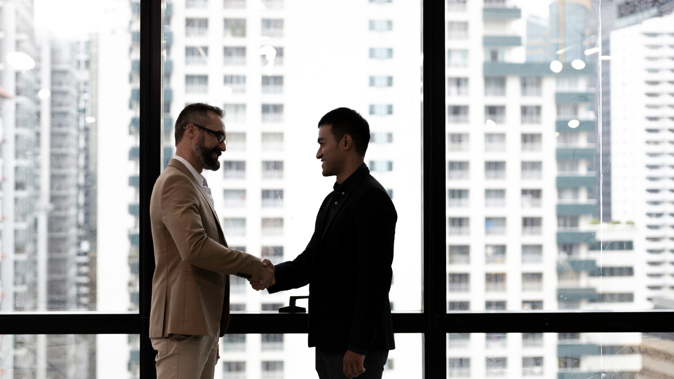 Two men shaking hands in front of a window overlooking a cityscape. One man wears a beige suit, the other a black jacket.