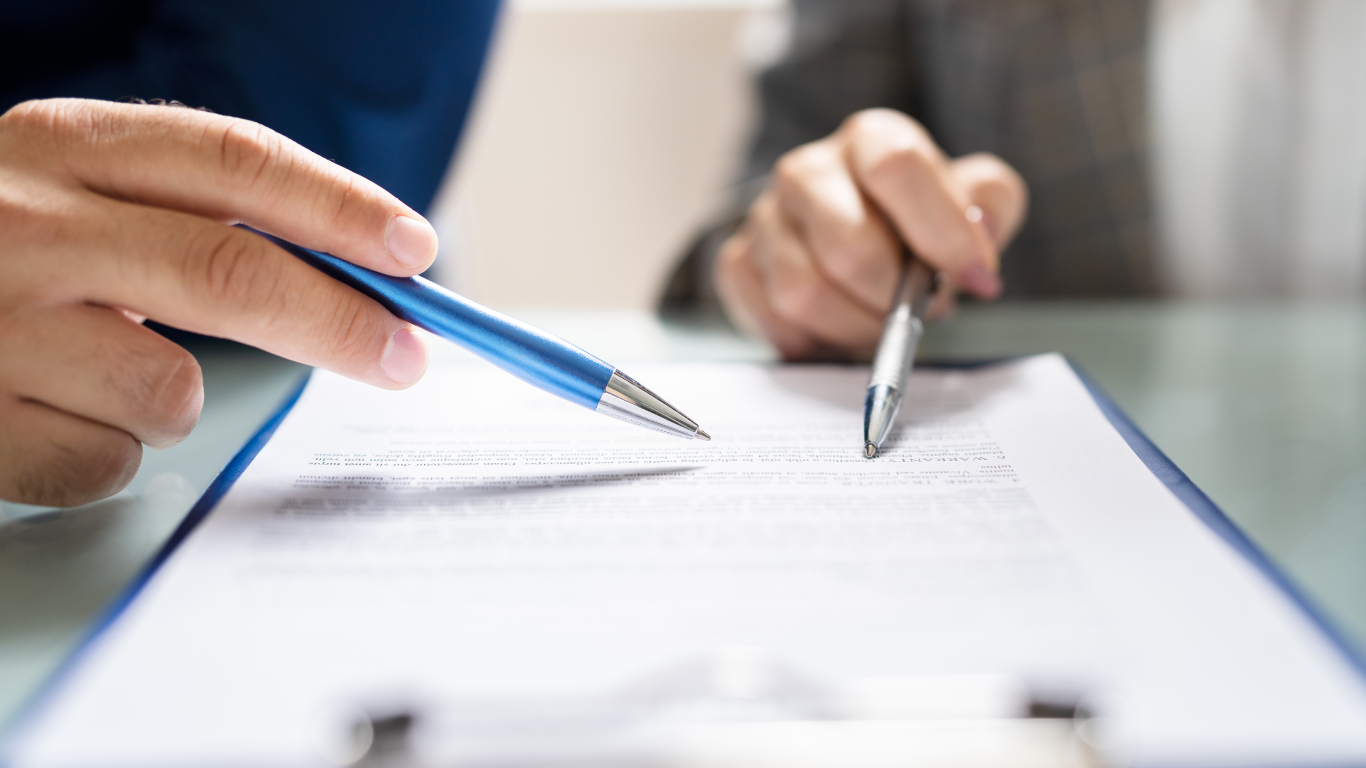 Two people pointing pens at a document on a clipboard.