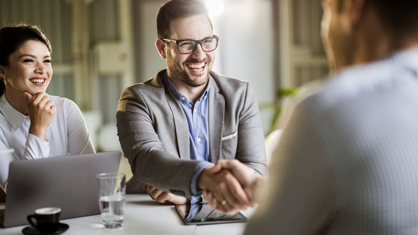 People in a business meeting, shaking hands. Smiling, with a laptop, coffee, and glass of water on the table.
