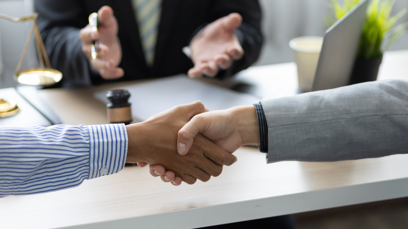 Two people shaking hands in an office, legal scales and a laptop on desk.
