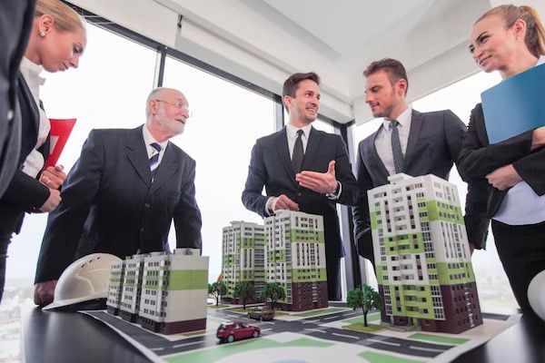 Businesspeople in suits gather around a model of buildings, discussing a project in an office.