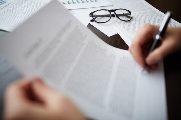 Person's hands writing on a document with a pen. Glasses and other papers are on the desk nearby.