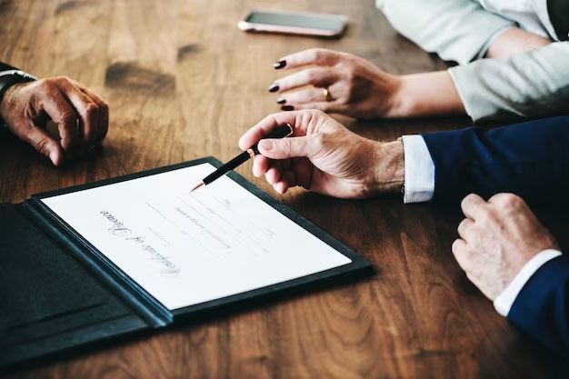 Hands signing a document in a black folder on a wooden table, two other people present with a phone.
