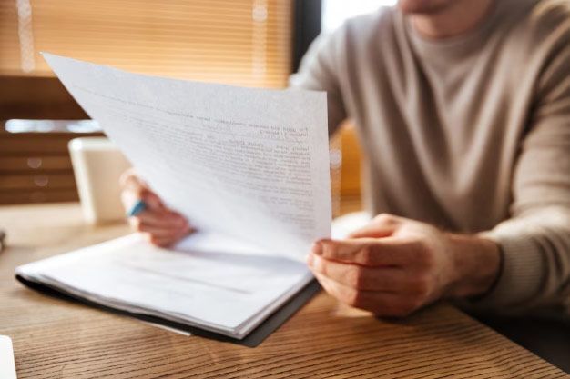 Person reviewing documents at a wooden table, holding a sheet and notebook.