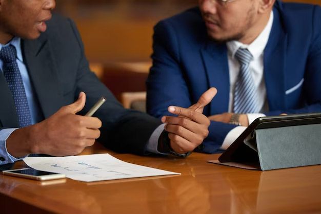 Two men in suits gesturing and discussing documents at a table.