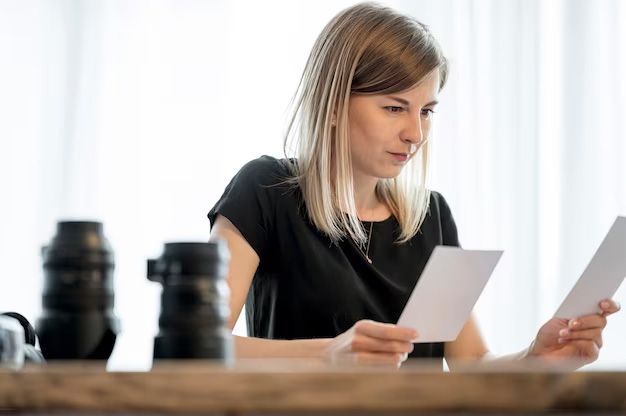 Woman looking at photos, seated at a table with camera lenses in front of a window.