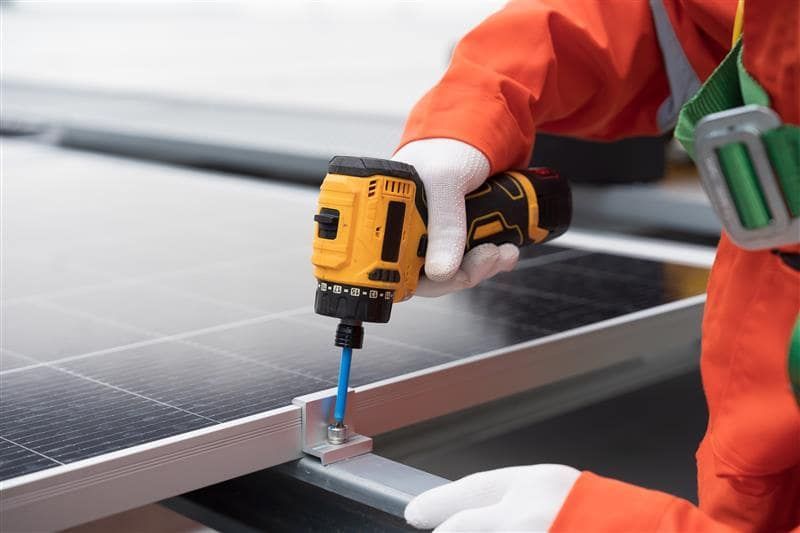 A worker in orange coveralls uses a yellow drill to install a solar panel.