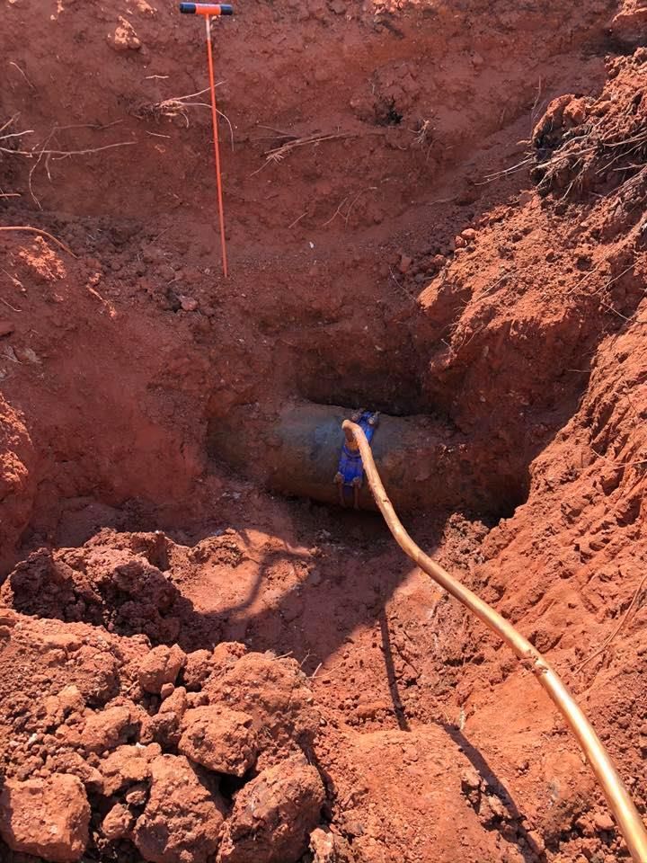 A pipe is exposed in a hole dug in red soil, with a probe and a hose nearby.