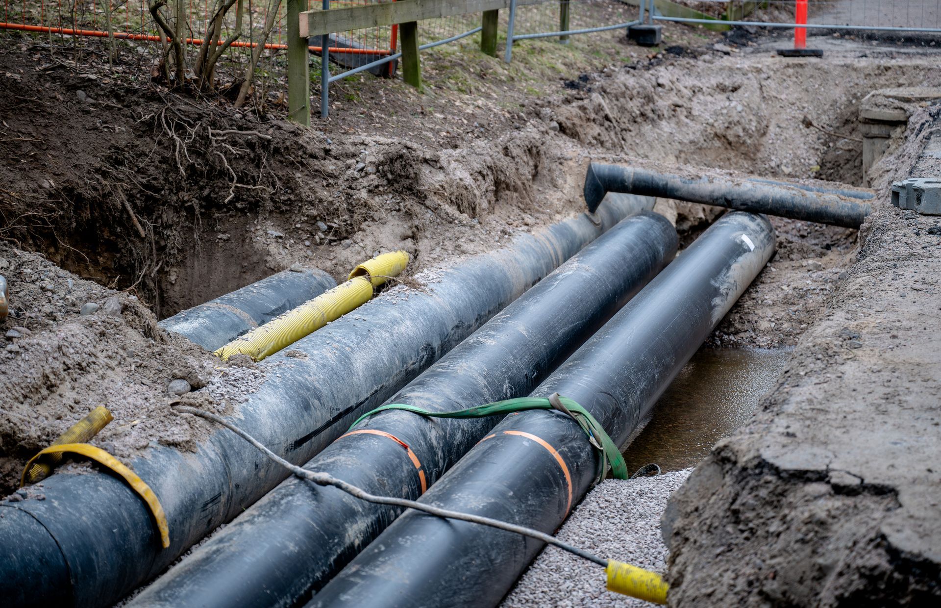 Black and gray pipes in an open trench, likely for utilities, set in dirt and gravel, outdoors.
