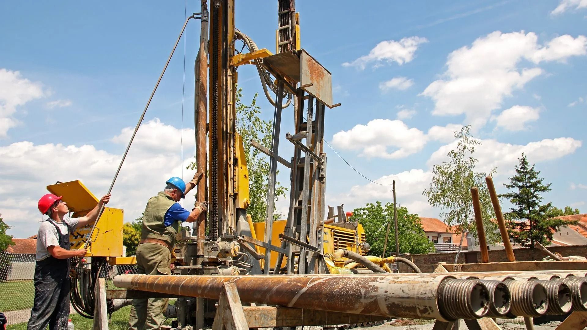 Two workers operate a yellow drilling rig outdoors on a sunny day.