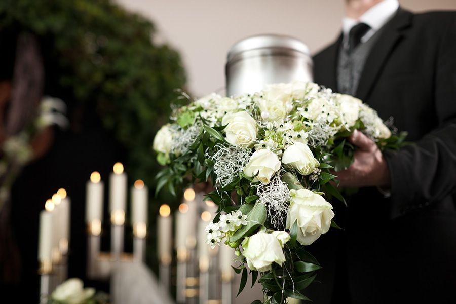 Person in black suit holding an urn with white flowers at a funeral, candles lit in background.