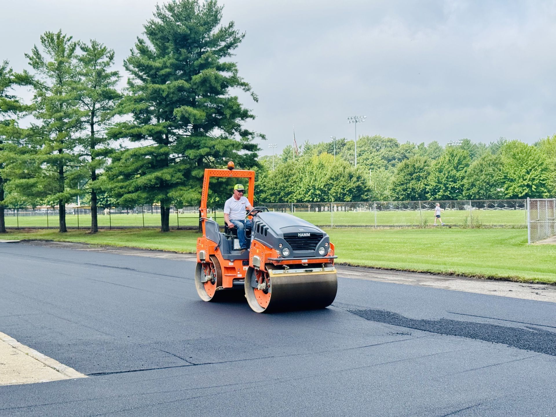 Man operating an orange asphalt roller on a newly paved road. Trees and sky in the background.