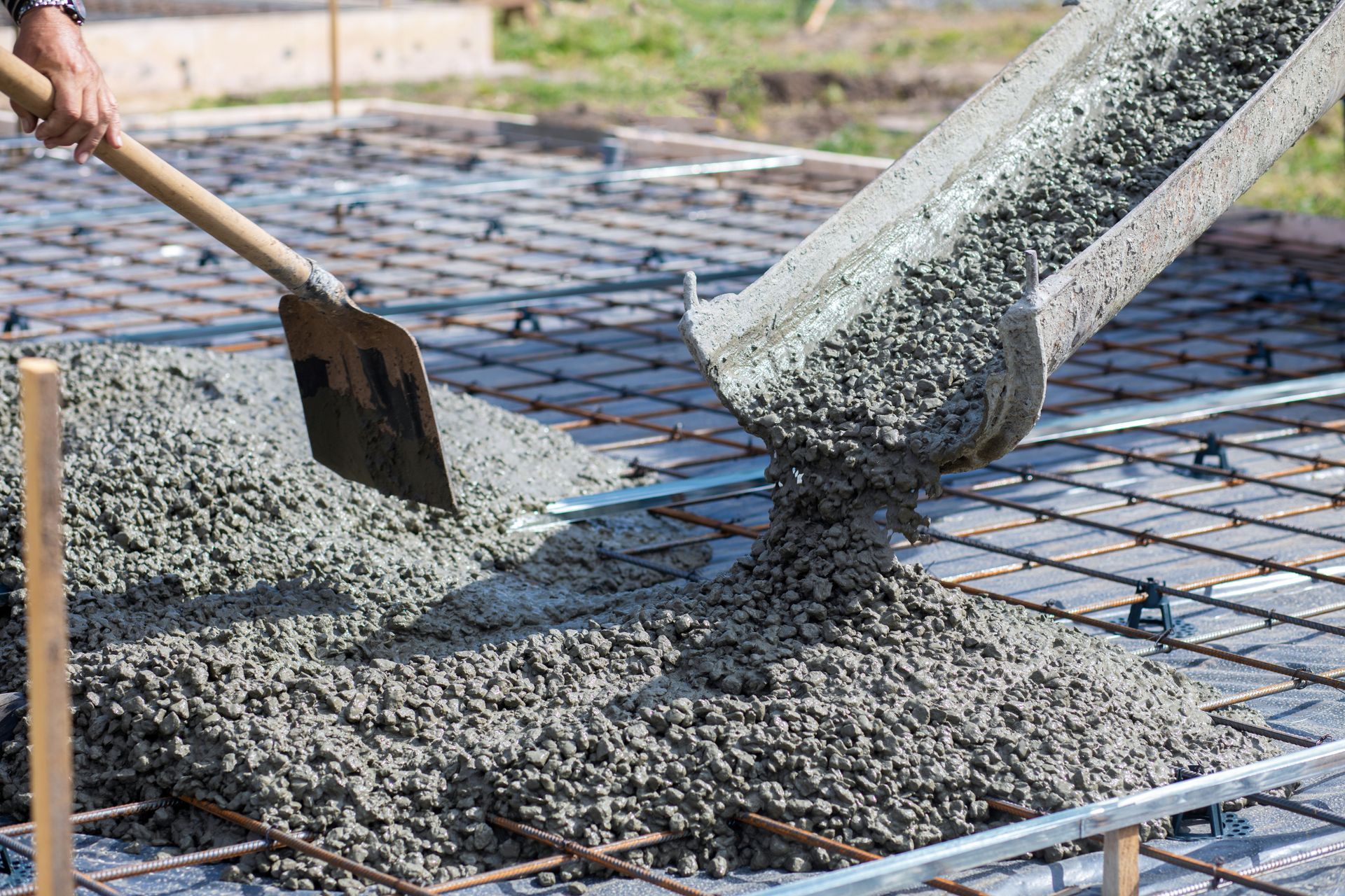 Pouring wet concrete onto a reinforced steel grid, with a shovel nearby, outdoors.