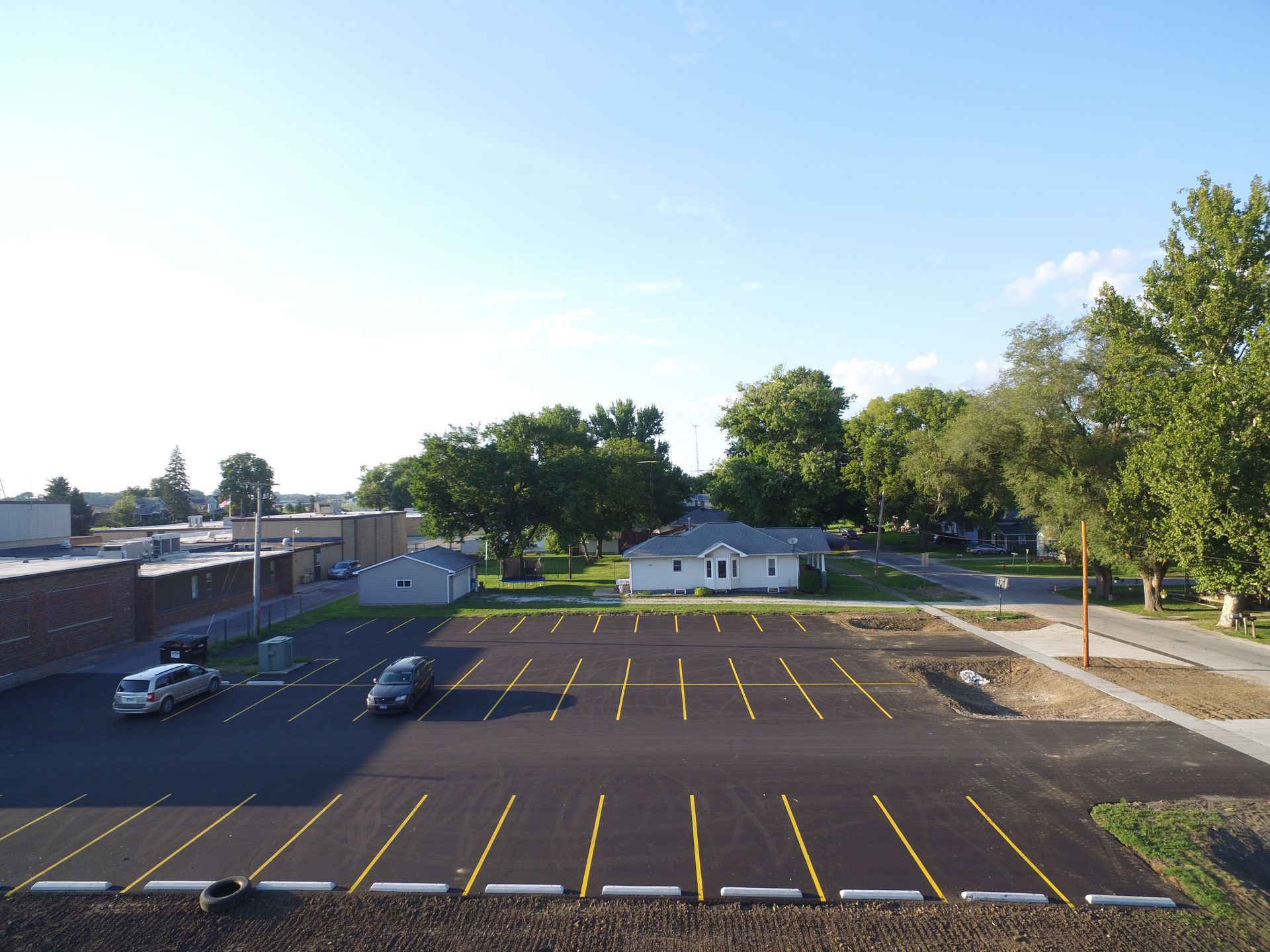 Empty asphalt parking lot with yellow parking space lines, small buildings, and trees.