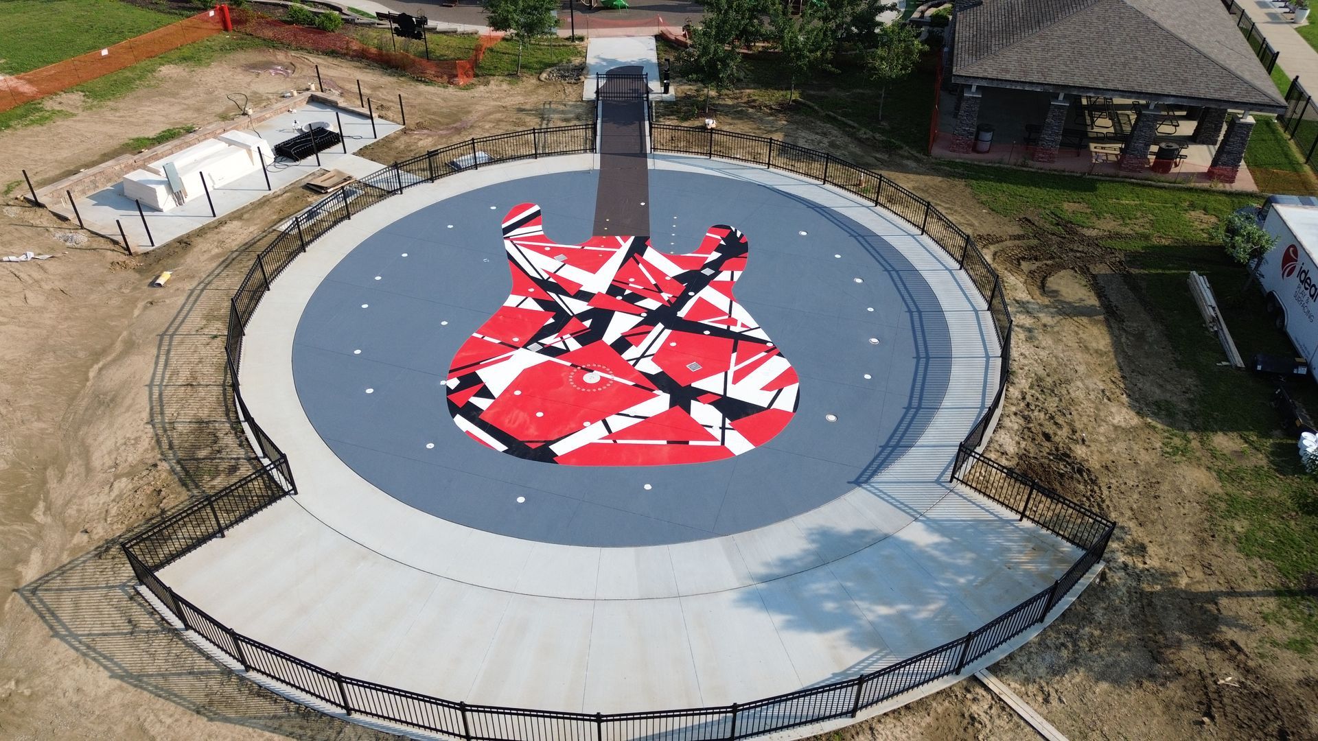 Aerial view: large guitar-shaped artwork in red, black, and white on a circular platform, outdoor setting.