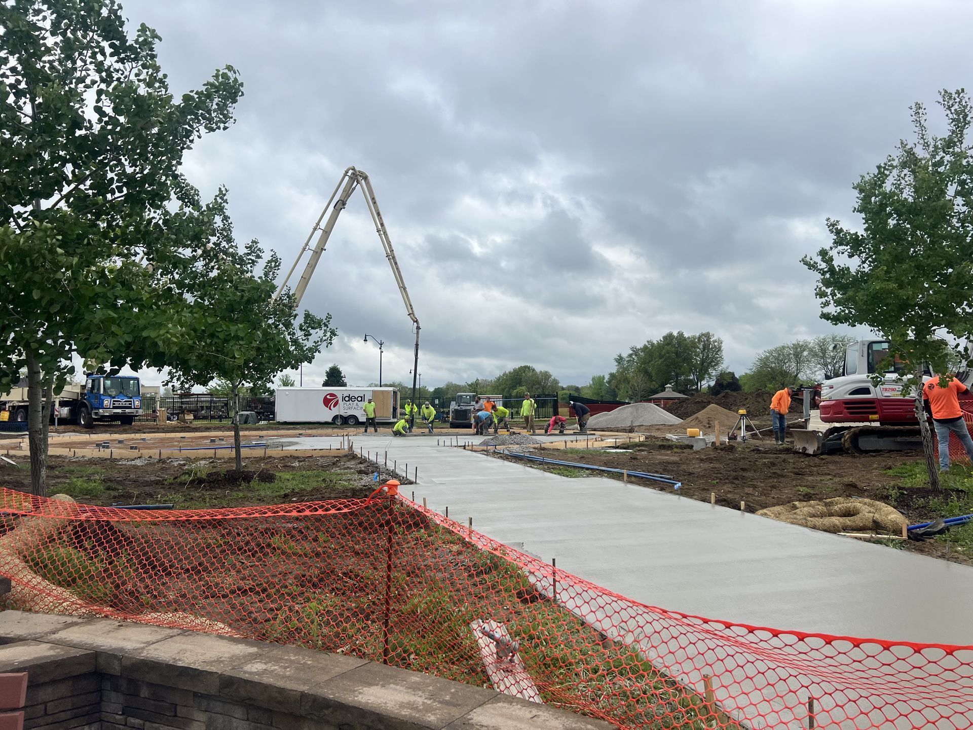 Construction site with a concrete path being poured. Workers in safety vests surround equipment under a cloudy sky.