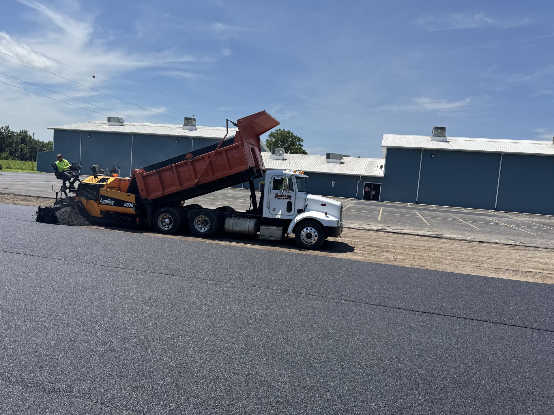 Asphalt paving: Dump truck unloading onto a paver, laying fresh asphalt on a sunny day.