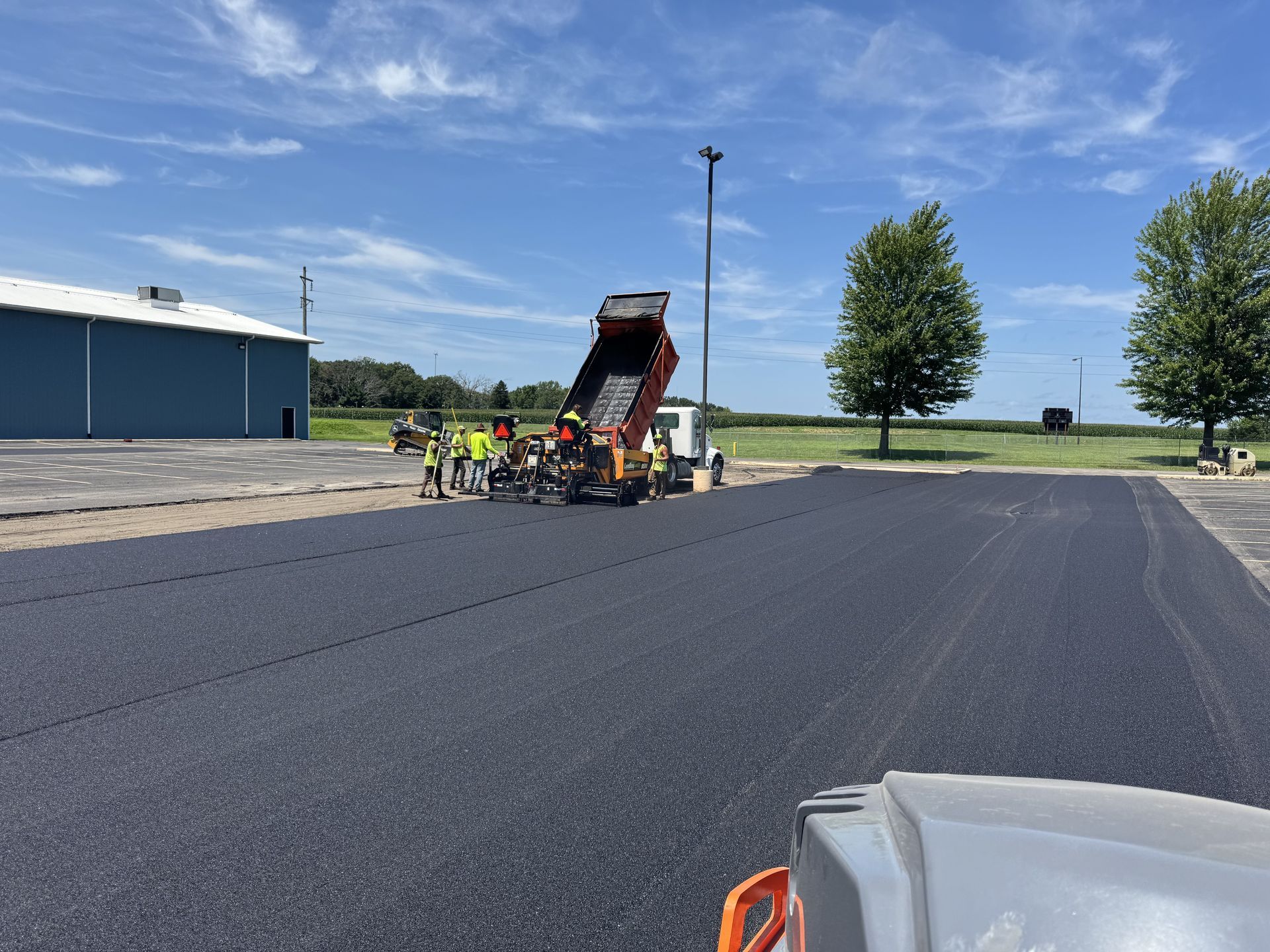 Asphalt paving crew at work, a dump truck unloading into a paving machine. Blue building and sky.