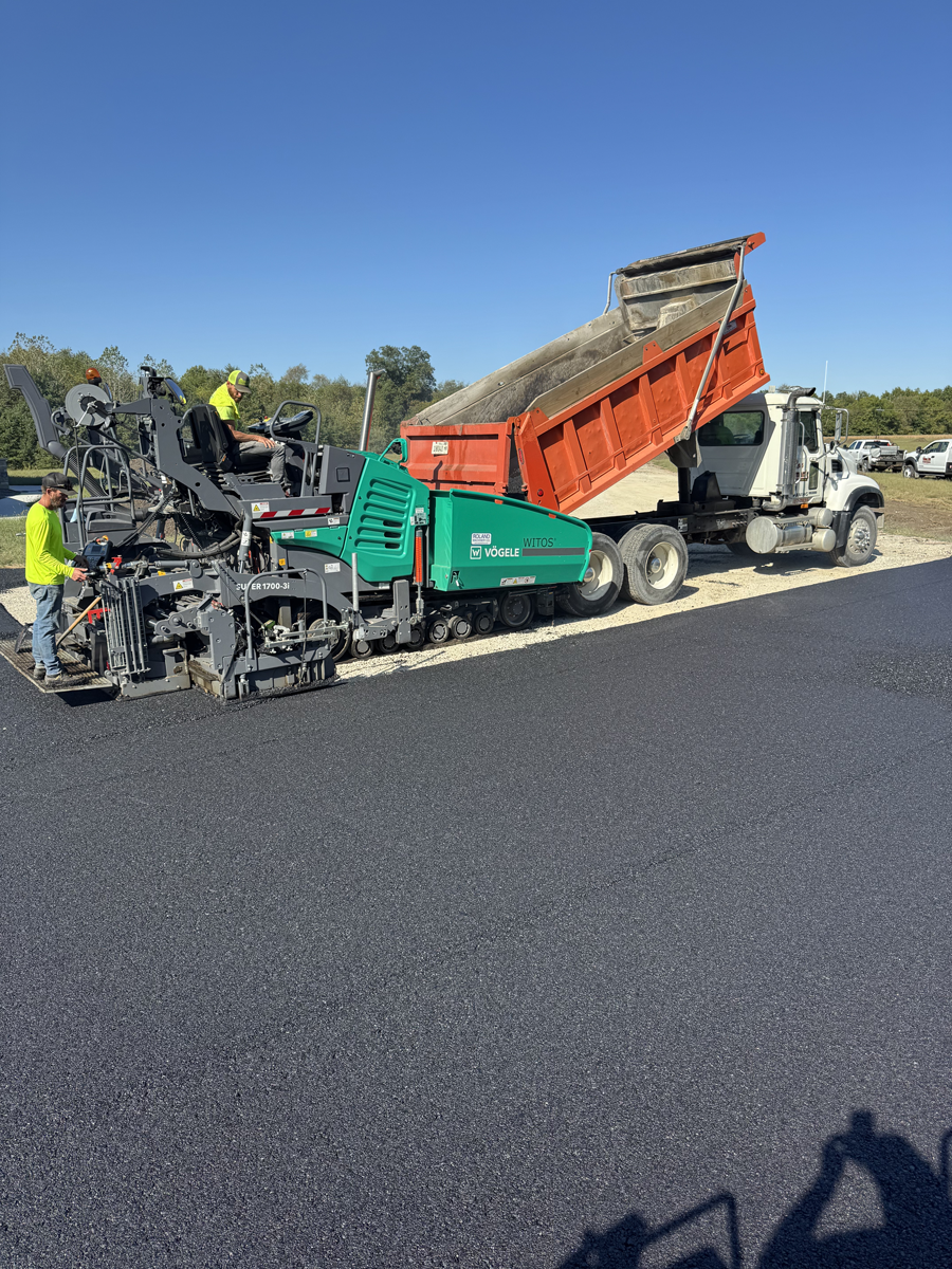 Asphalt paving operation: dump truck unloading asphalt into a paving machine on a sunny day.
