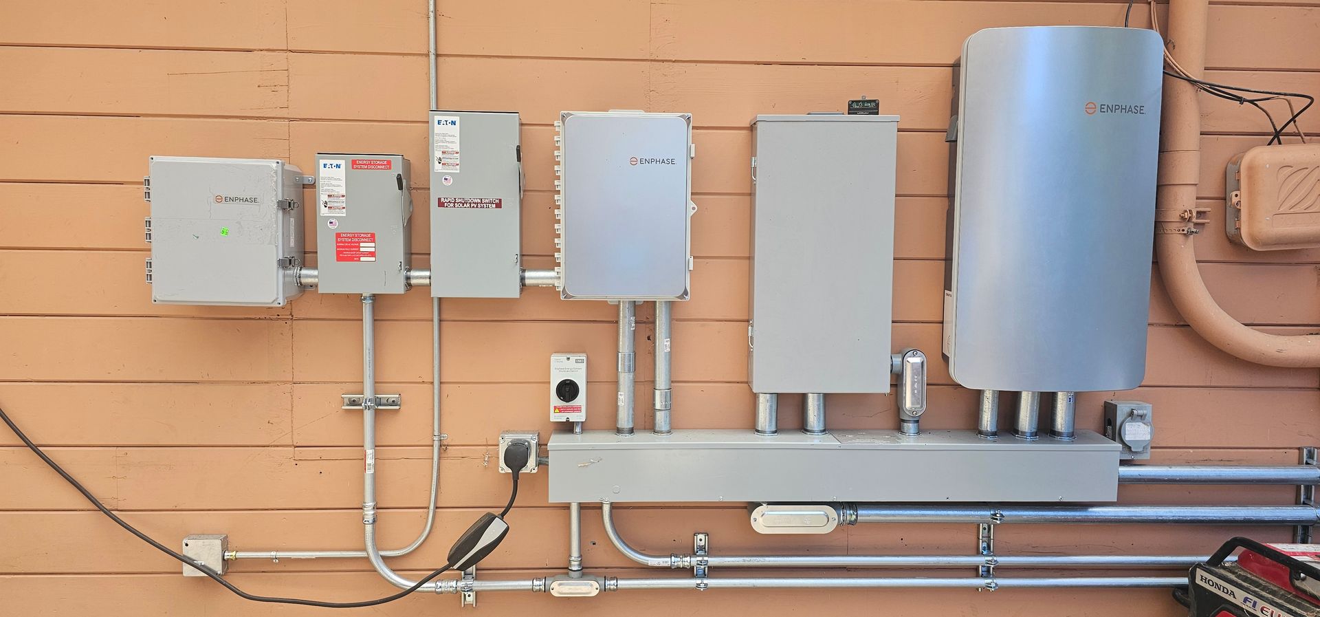 Row of electrical and utility control boxes mounted on a tan exterior wall, with conduit and piping below.