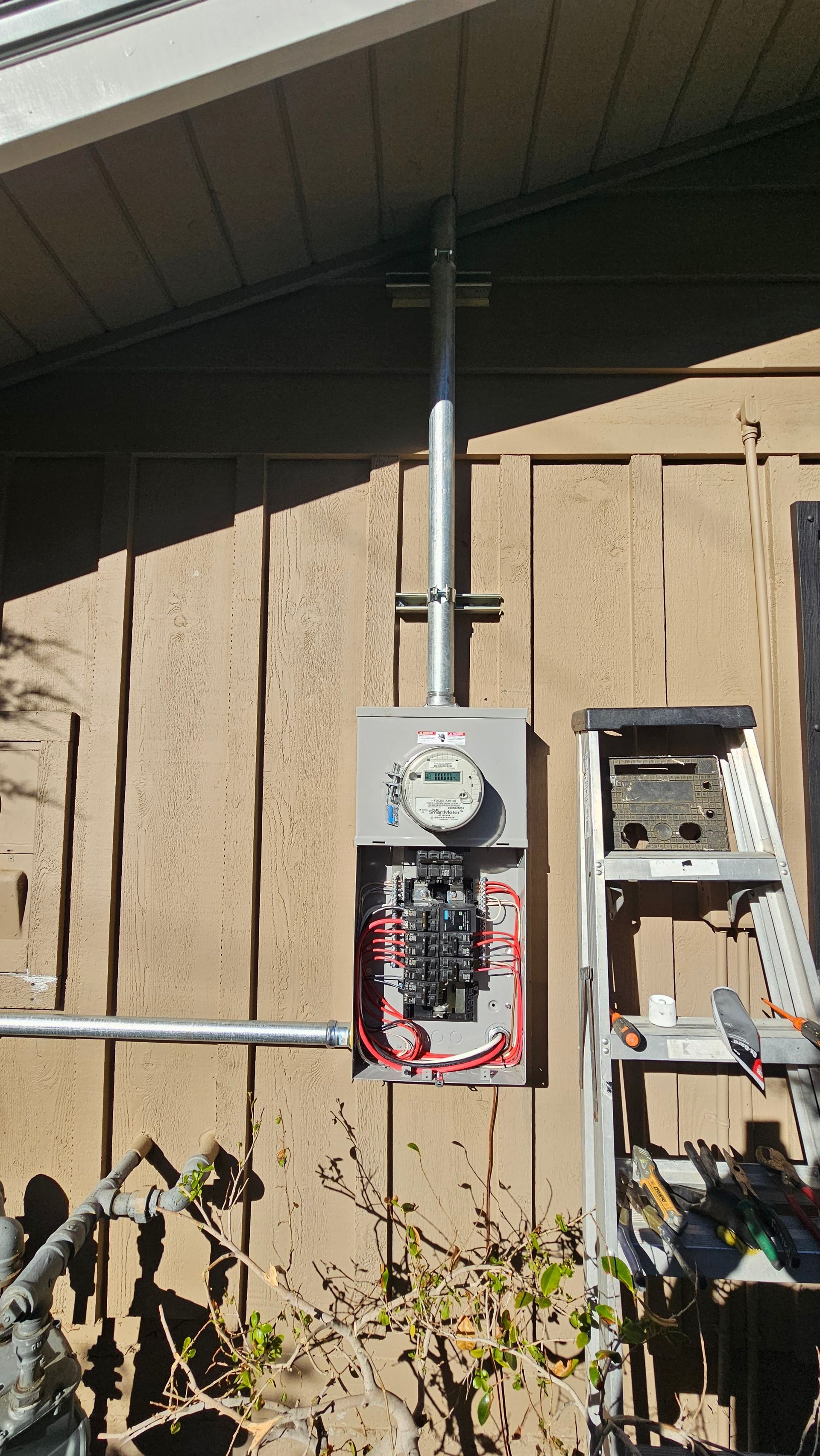Electric meter and open breaker panel mounted on tan exterior wall under a roof overhang.