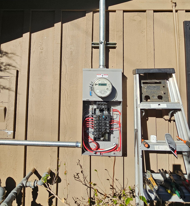 Open electrical meter and wiring panel mounted on a wooden exterior wall.