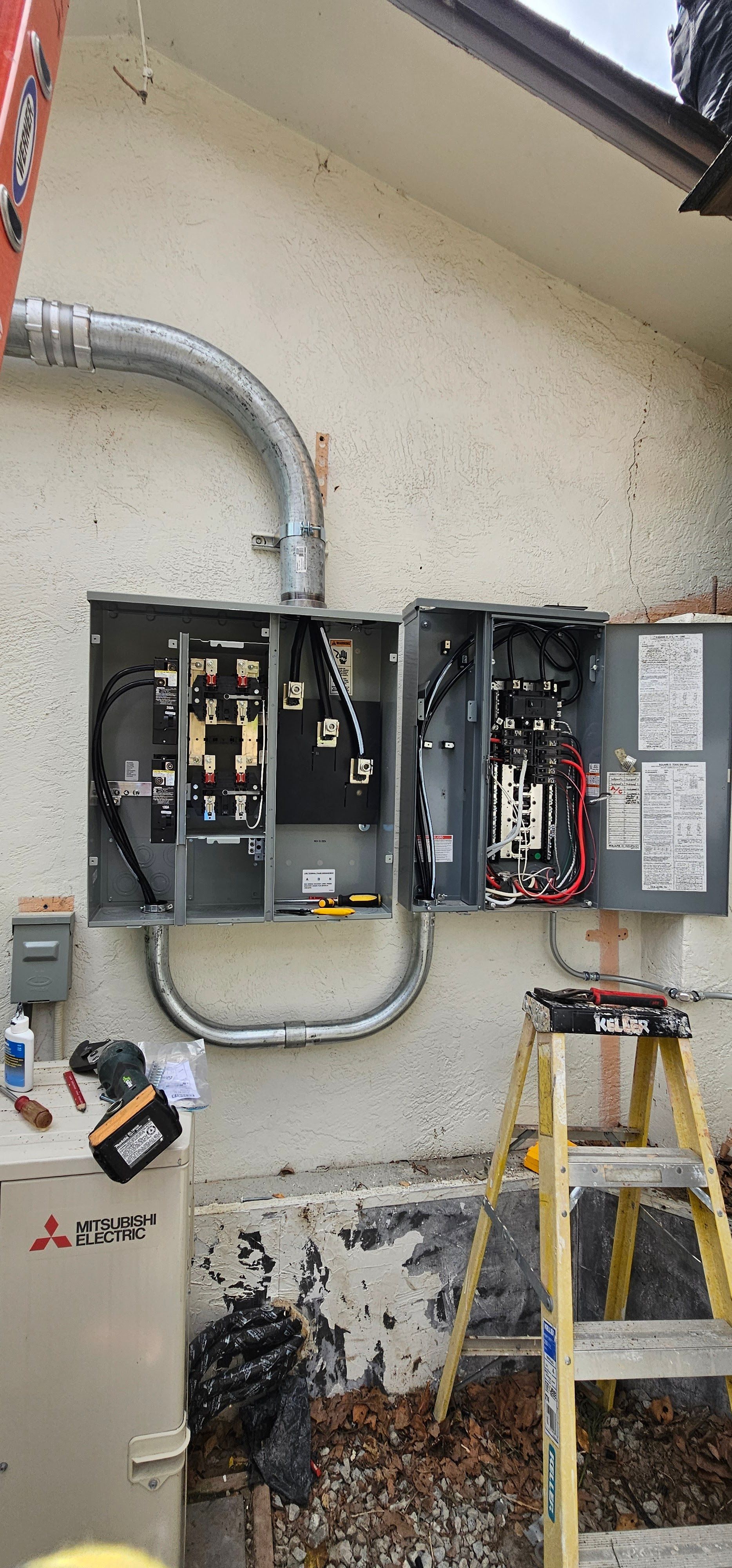 Open electrical panels on an exterior wall with conduit, ladder, and rusted debris below