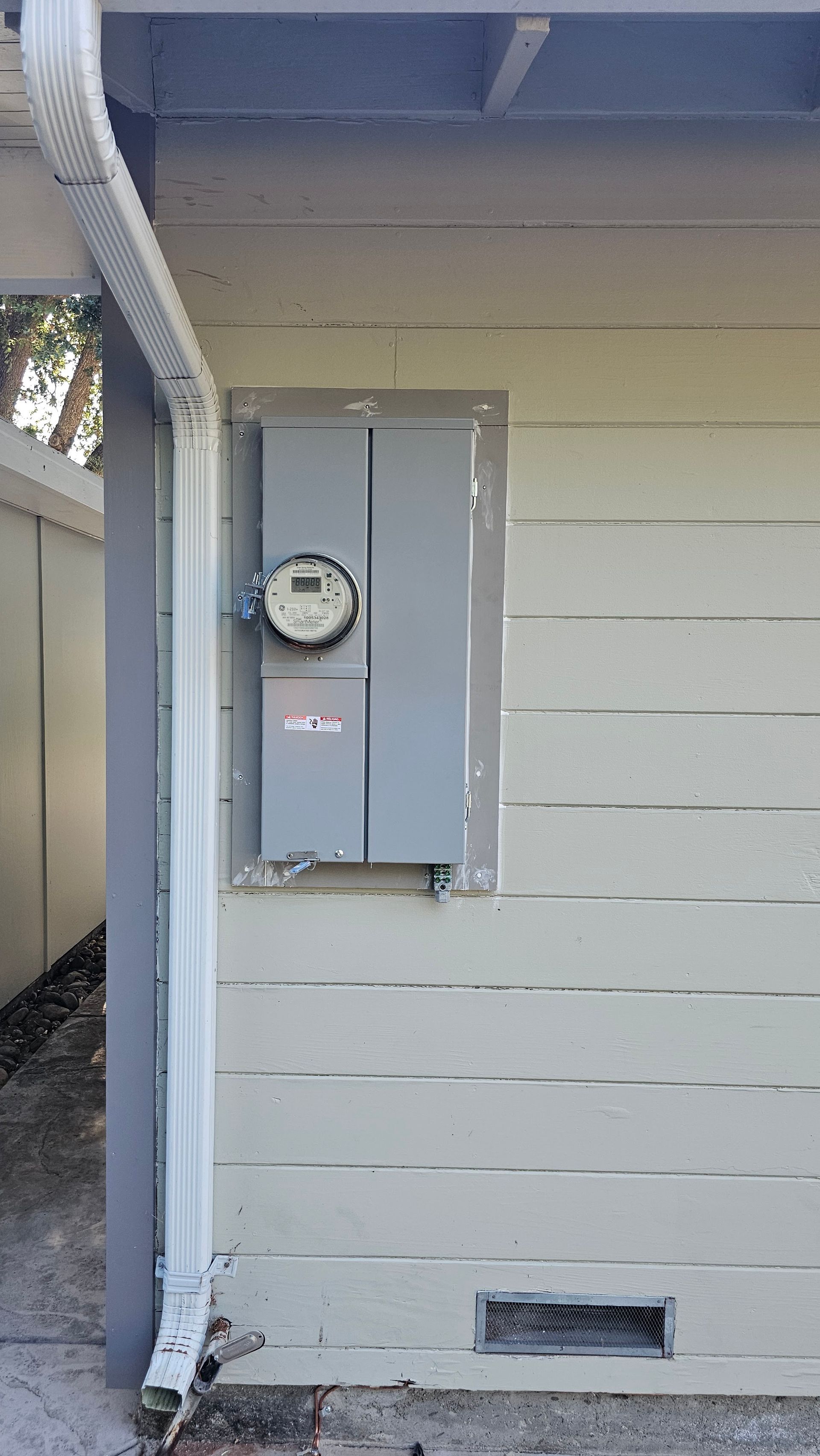 Outdoor electric meter box mounted on beige house siding under a roof overhang