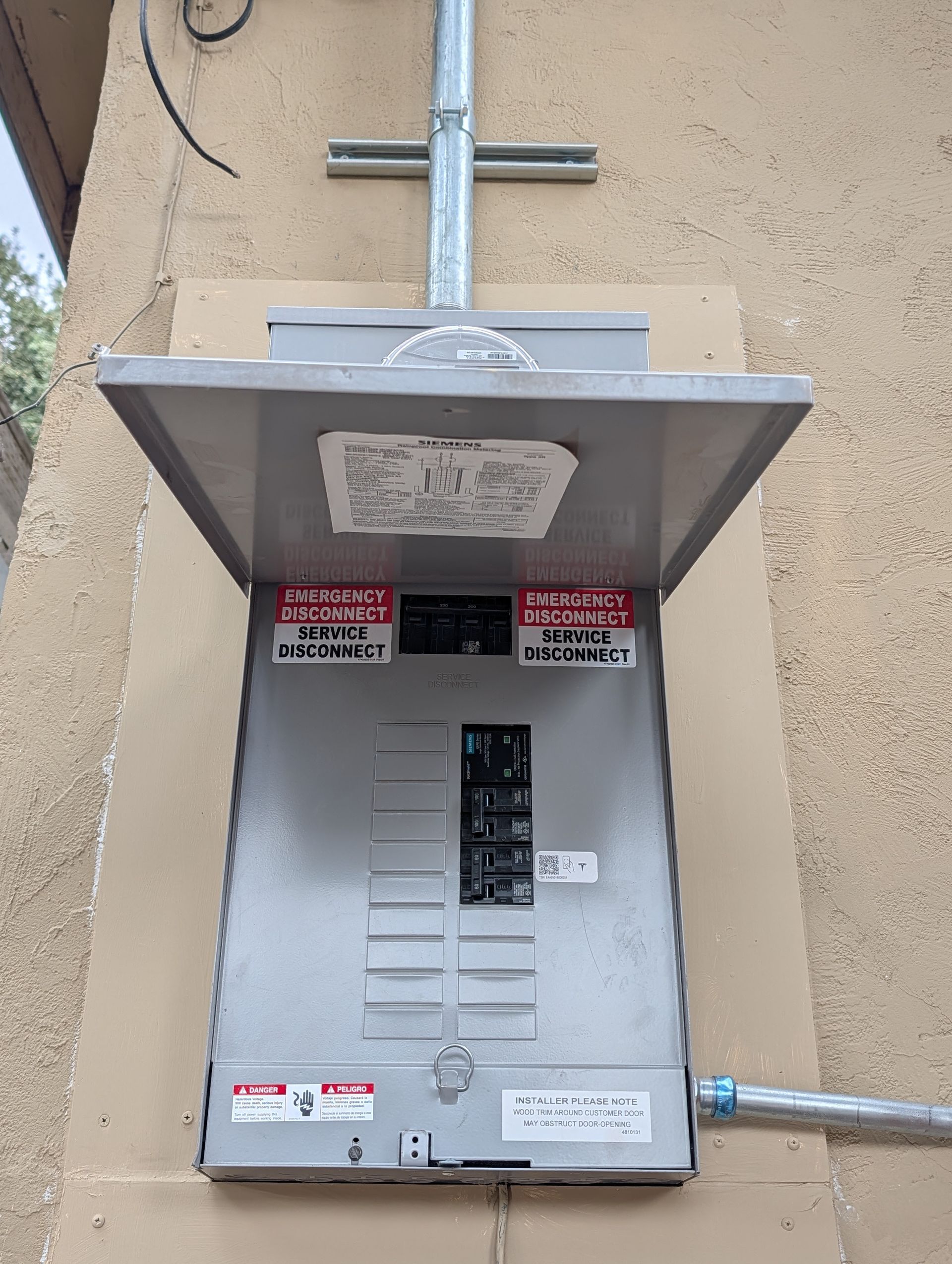Outdoor electrical breaker panel mounted on a beige wall, with metal conduit and warning labels.