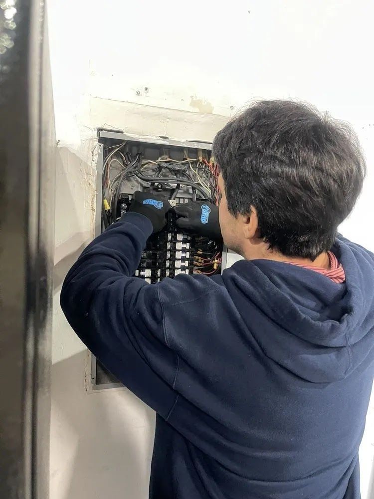 Person in a navy hoodie working inside an open electrical panel with gloves on.