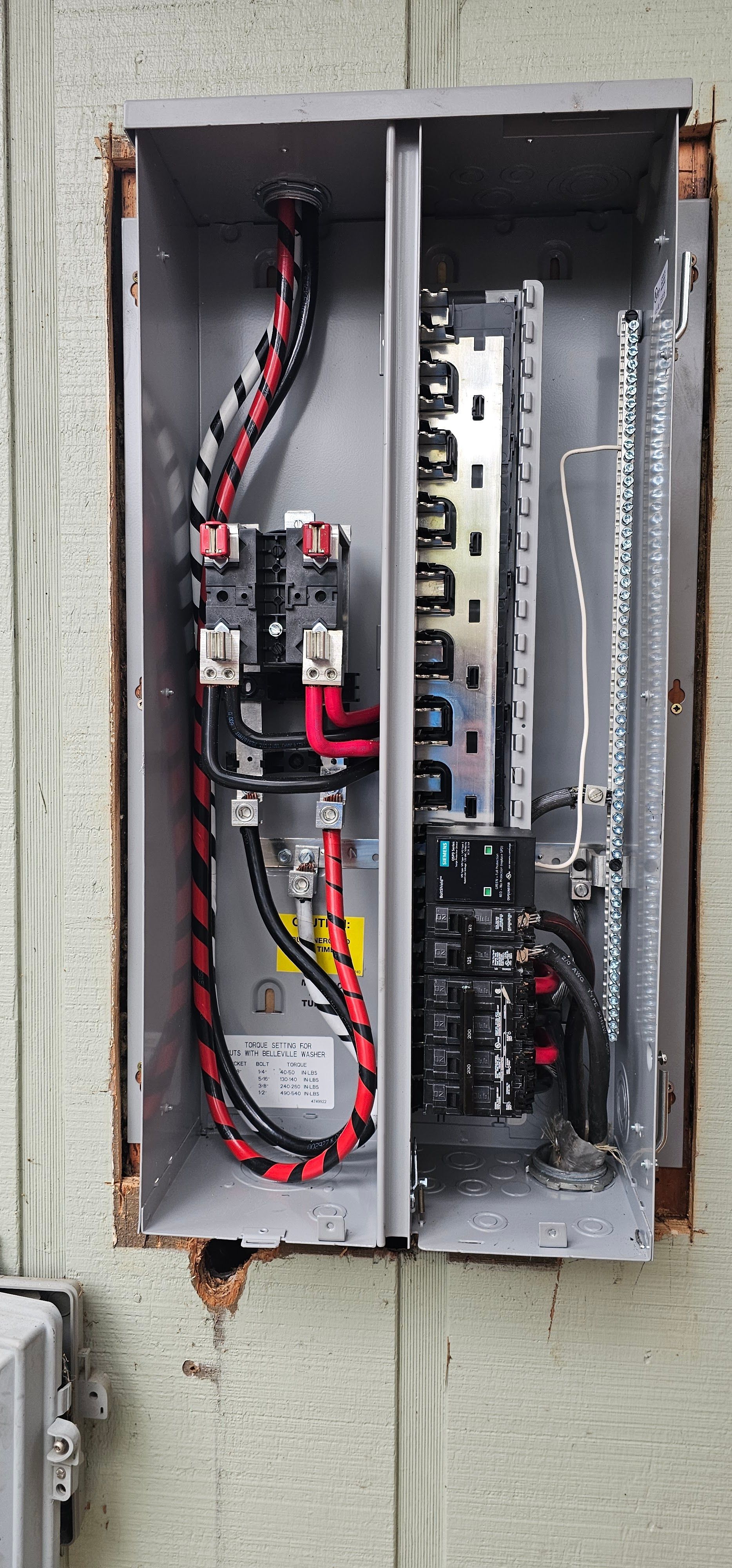 Open electrical breaker panel with exposed wires and circuit breakers mounted on a wall.