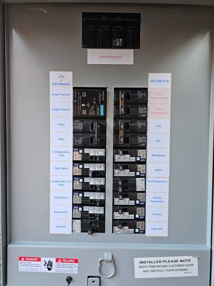 Open electrical breaker panel with labeled circuits and warning labels on a gray wall.