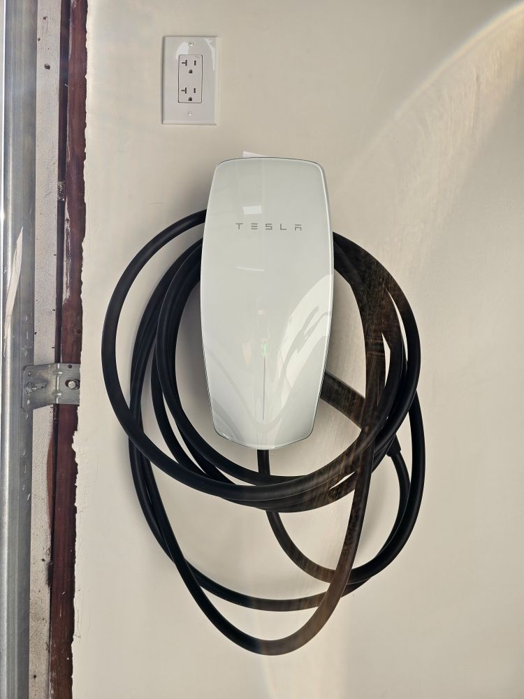 White wall-mounted Tesla EV charger with coiled black cable beside an outlet.