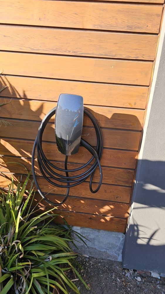 Outdoor wall-mounted cable reel on a wooden fence beside a house corner and plants
