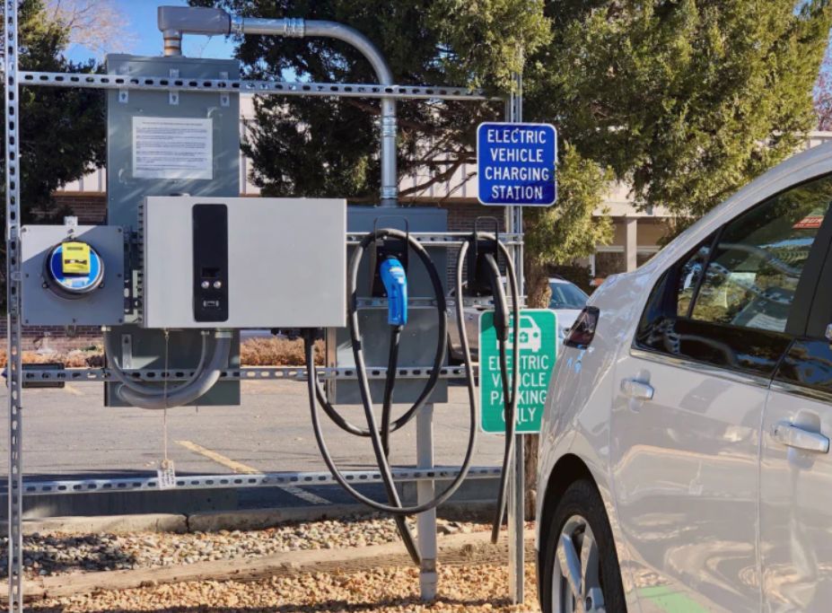 Electric vehicle charging station beside a parked white car, with blue and green signs outdoors.