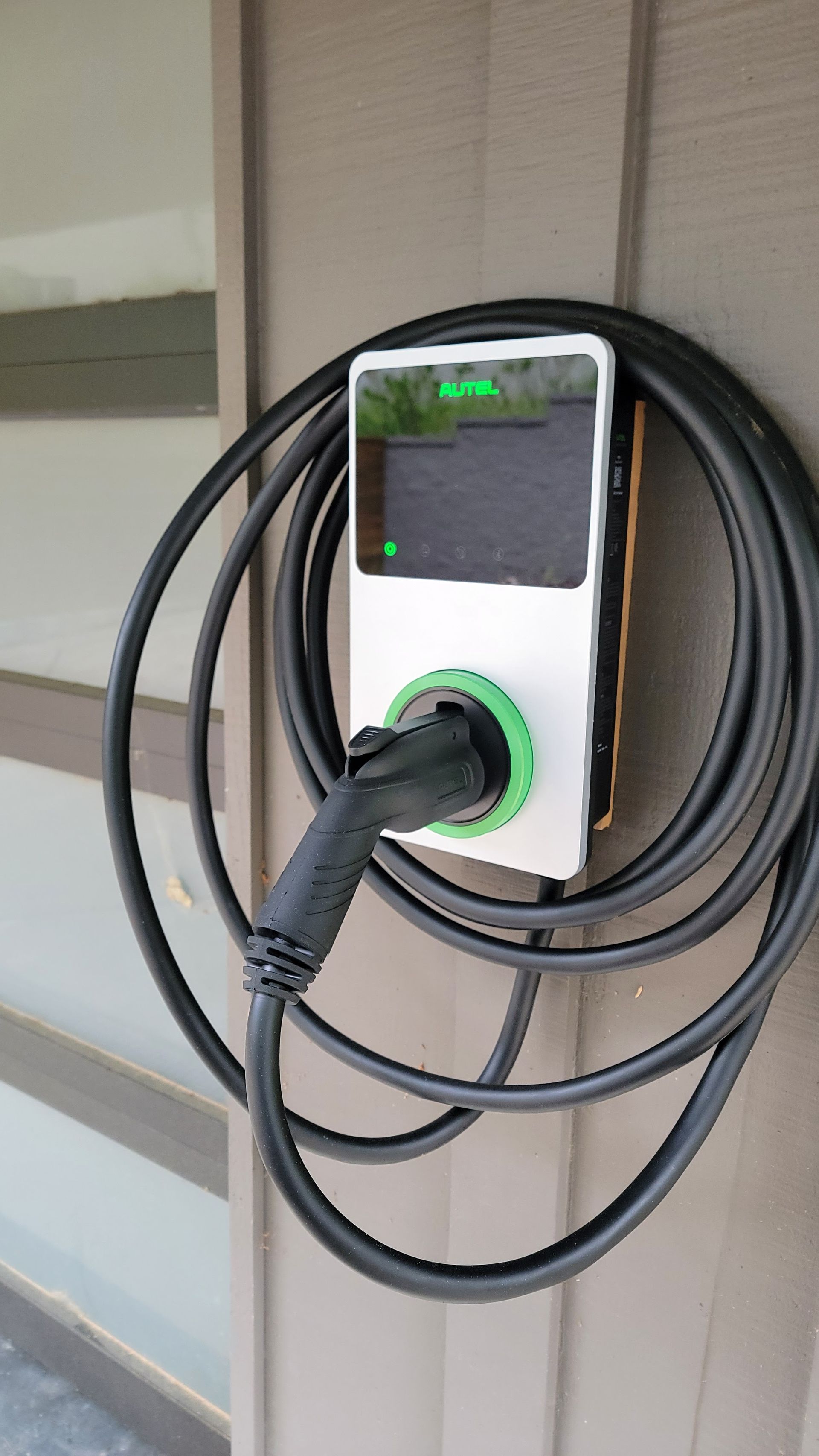 Wall-mounted EV charger with coiled black cable and lit green charging ring outdoors