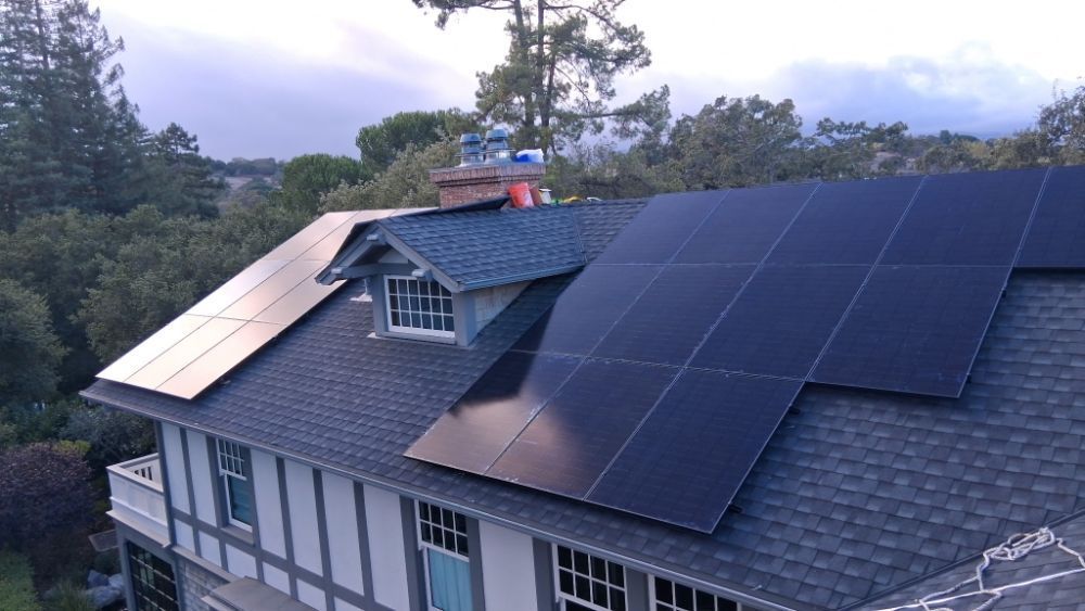 Solar panels mounted on a house roof at dusk, with trees and cloudy sky in the background.