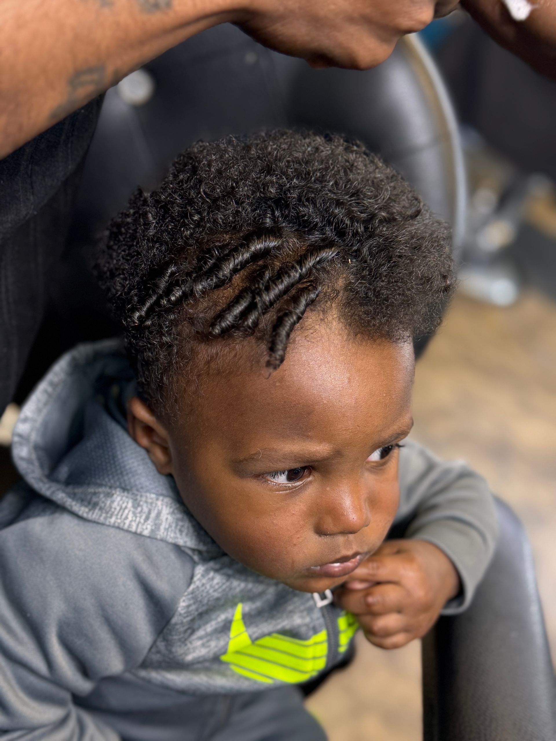 A little boy with braids in his hair is sitting in a chair.