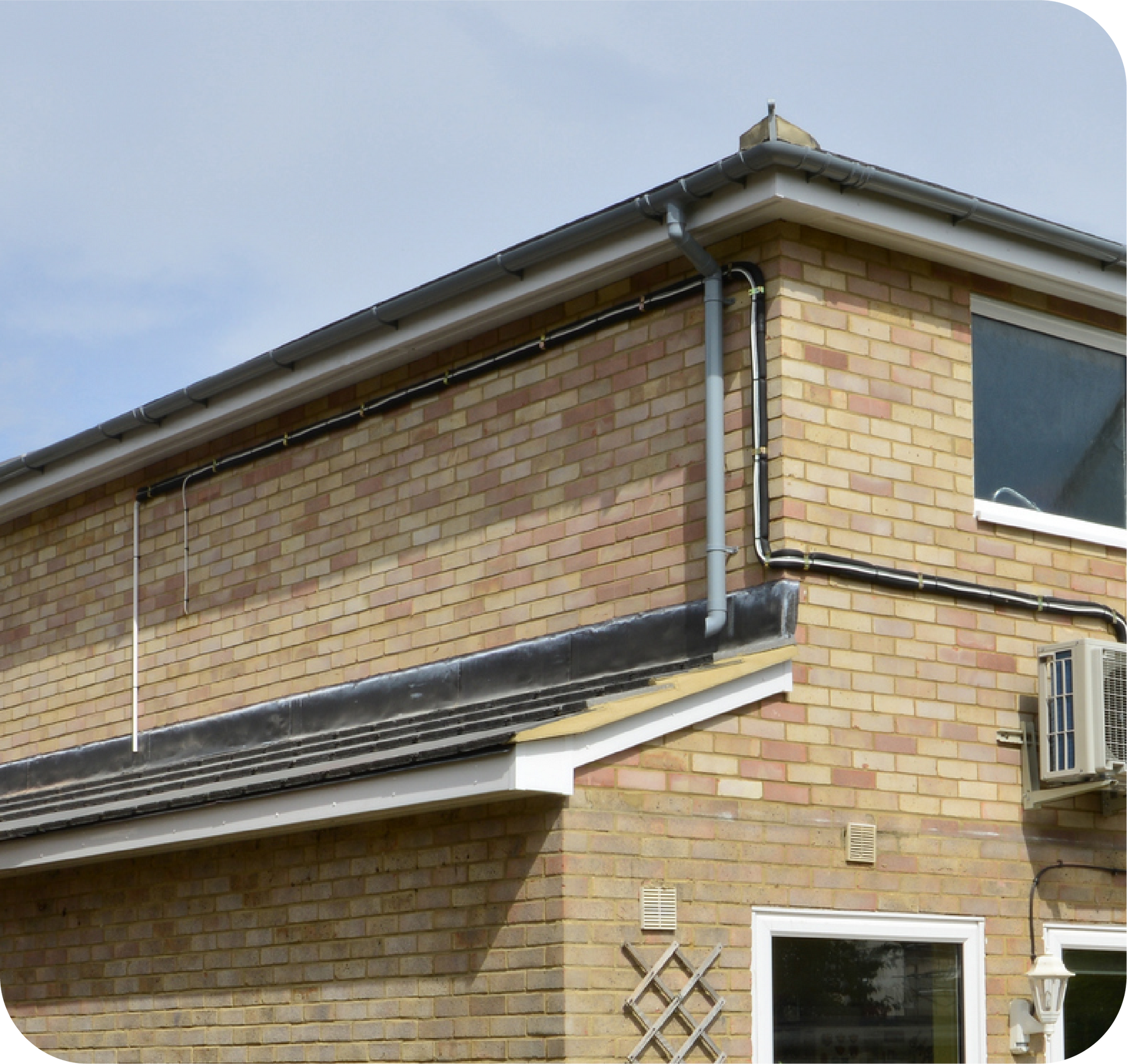 Corner of a brick building with a dark roof and metal guttering against a partly cloudy sky.