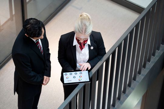 Two people in suits looking at a tablet near a staircase railing.