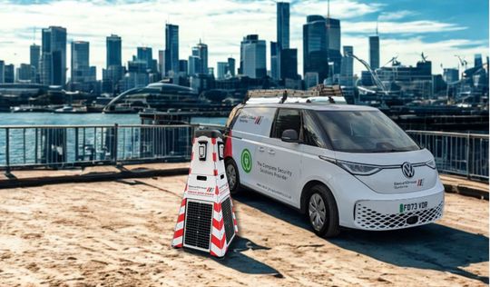 White van with surveying equipment, city skyline in the background, near water.