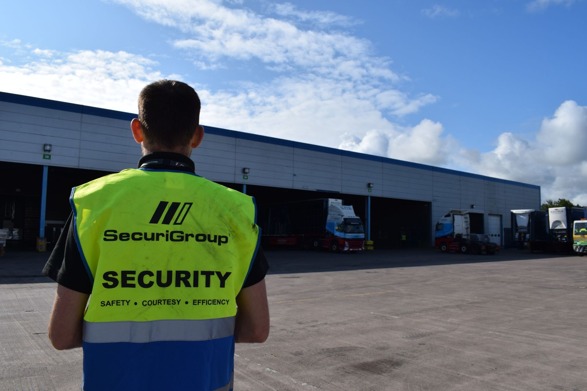 Security guard in a yellow vest, standing in front of a warehouse with trucks, under a blue sky.