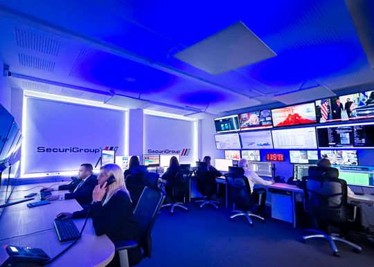 Security control room with staff at desks, monitors displaying feeds, under blue lighting.