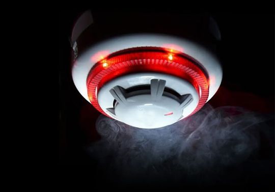 Smoke detector with red lights activated, emitting smoke in a dark room.