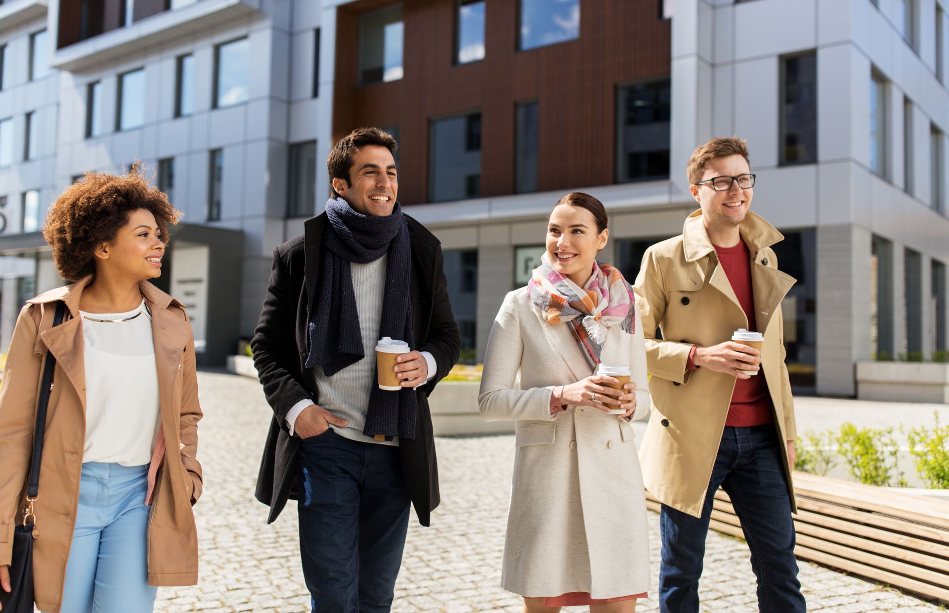 Four diverse young people walking and talking outside modern buildings, holding coffee cups.